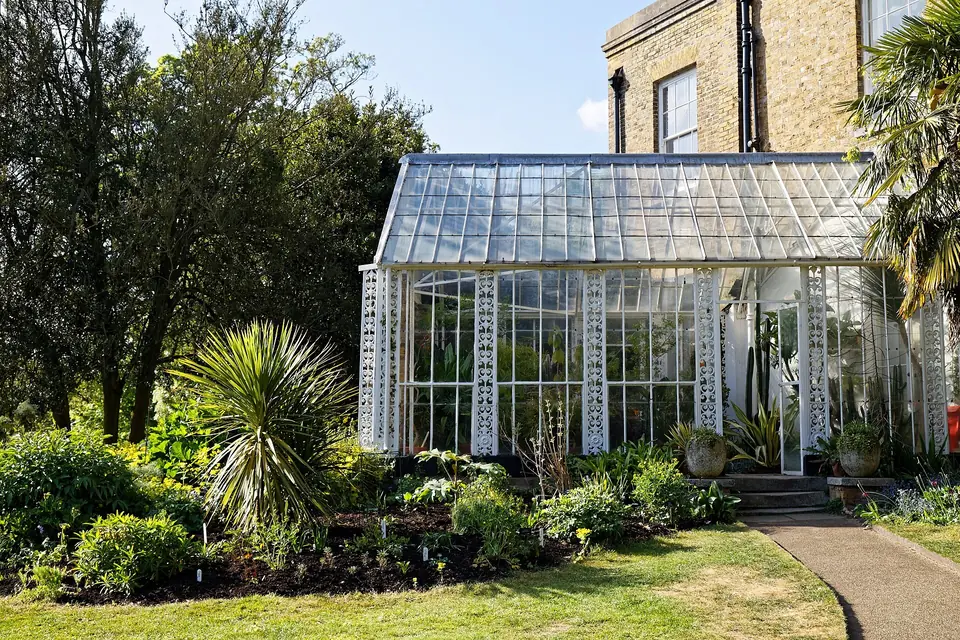 Véranda vitrée à structure blanche adossée à une maison en brique, entourée d'un jardin planté de palmiers et d'arbustes.